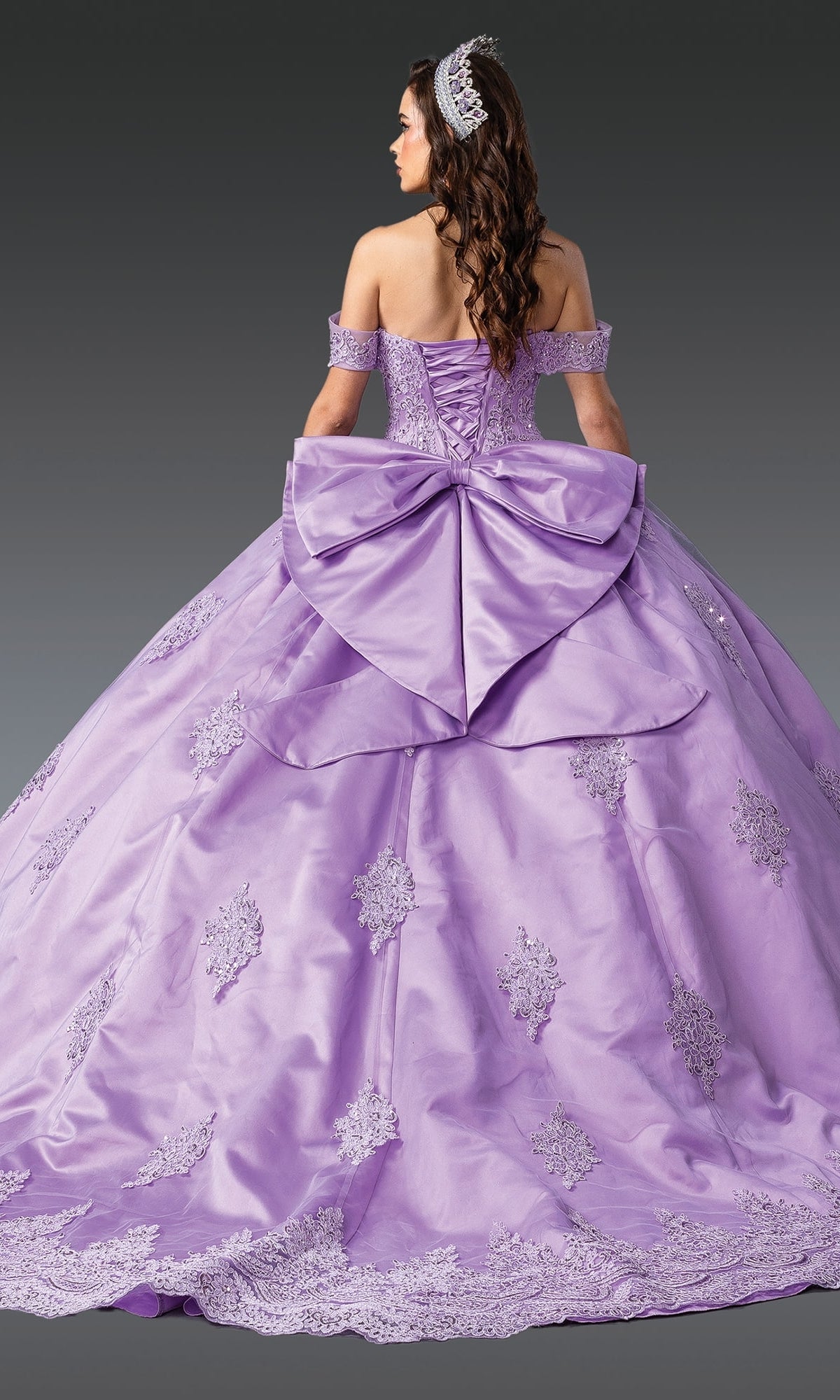 A woman faces away from the camera in the Quinceanera Dress 1913 By Dancing Queen, featuring lavender fabric, lace details, a corset bodice, and a large back bow against a gray background.