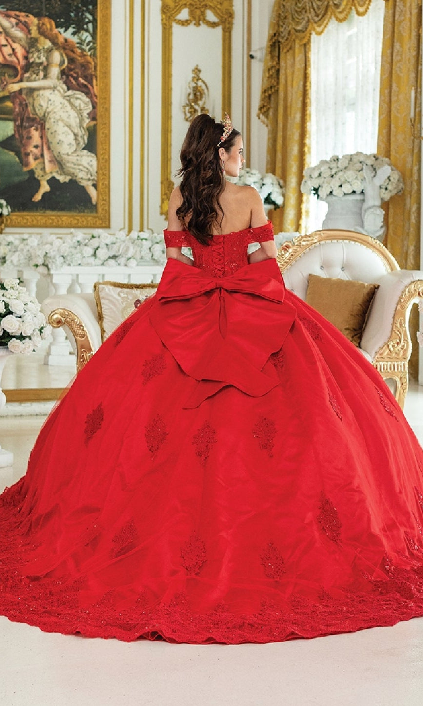 Wearing the Quinceanera Dress 1913 by Dancing Queen—a red off-the-shoulder ball gown with a corset bodice and large back bow—a woman sits on a white and gold sofa in an ornate, floral-adorned, gold-accented room.