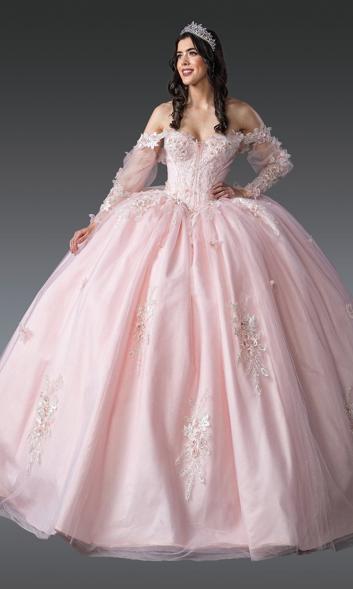 A woman smiles against a gray background, wearing the Quinceanera Dress 1914 By Dancing Queen—a pink gown with a corset bodice, lace details, and a tiara.