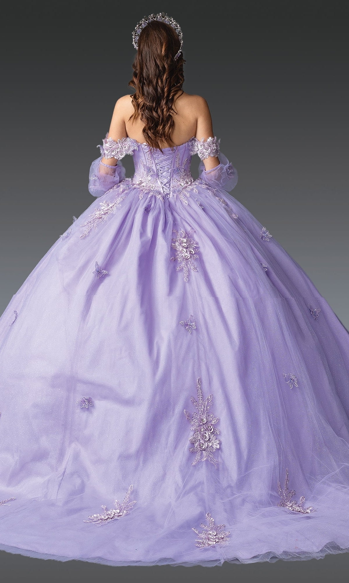 A woman with long brown hair wears the Quinceanera Dress 1914 By Dancing Queen, a lavender off-the-shoulder gown with a corset bodice and floral embroidery, seen from behind against a plain gray background.