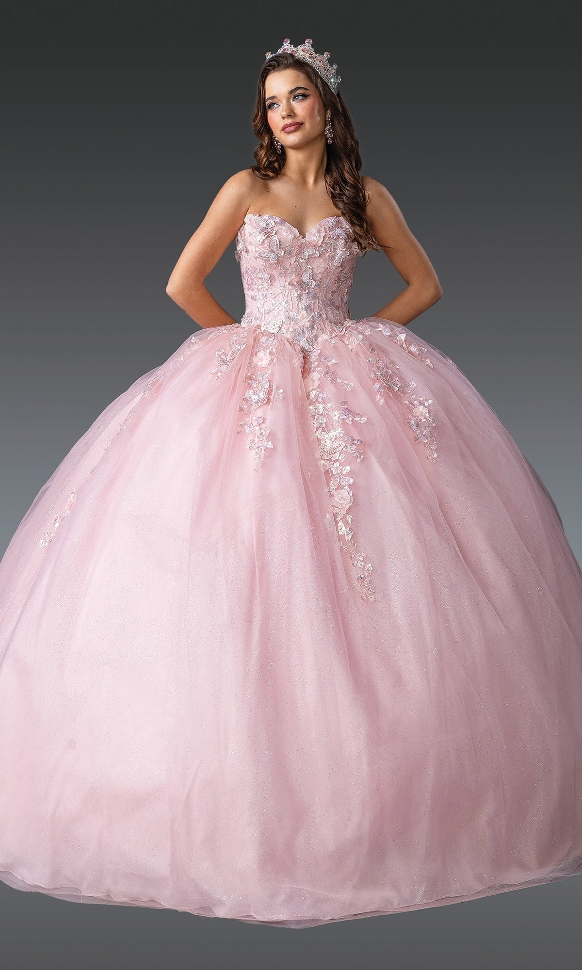 A young woman poses against a plain gray background in the Blush Pink Quinceanera Dress 1916 By Dancing Queen, featuring floral embellishments and a tiara.