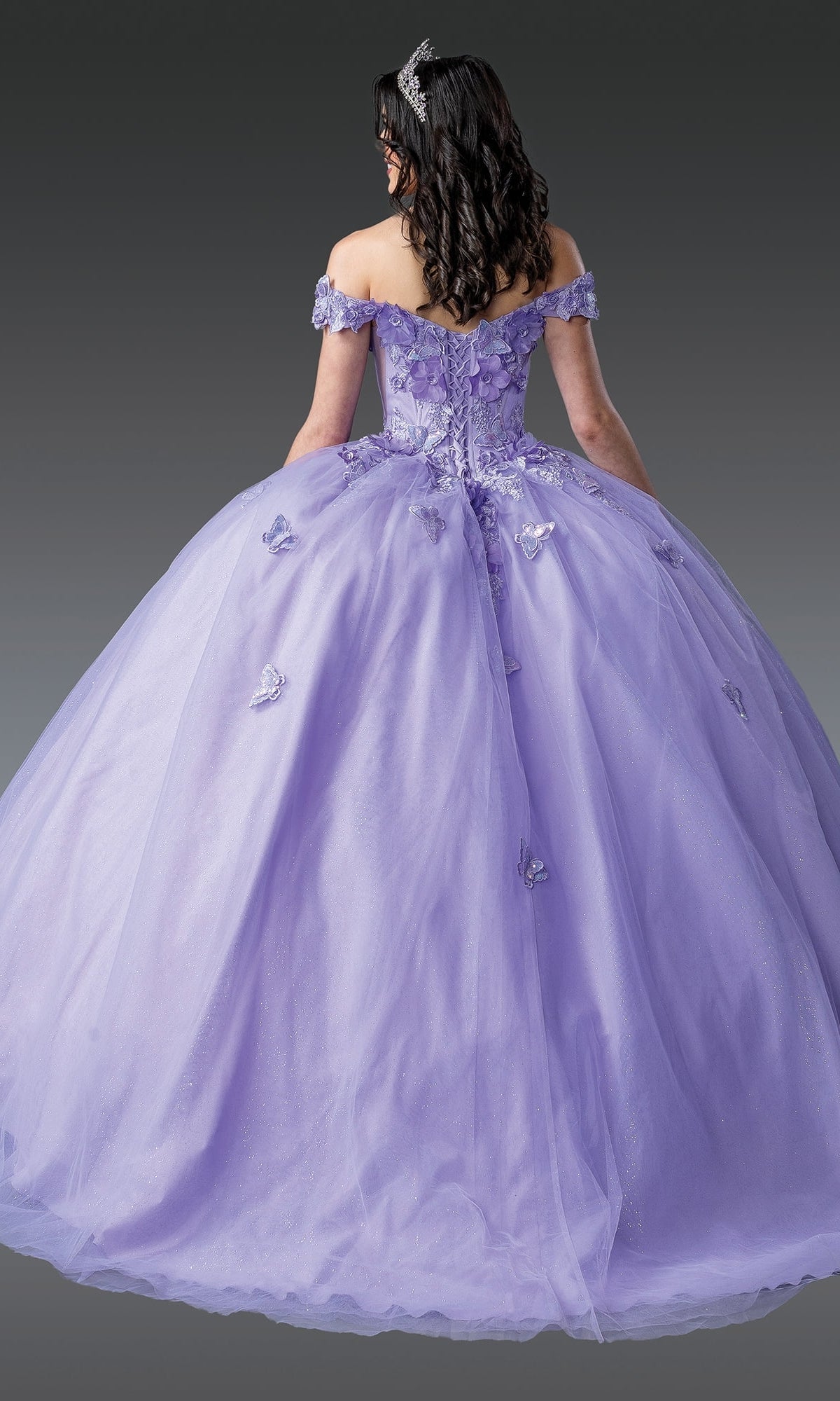 A woman faces away against a gray background, wearing the Quinceanera Dress 1917 by Dancing Queen—a floor-length, off-the-shoulder lilac gown with butterfly details.