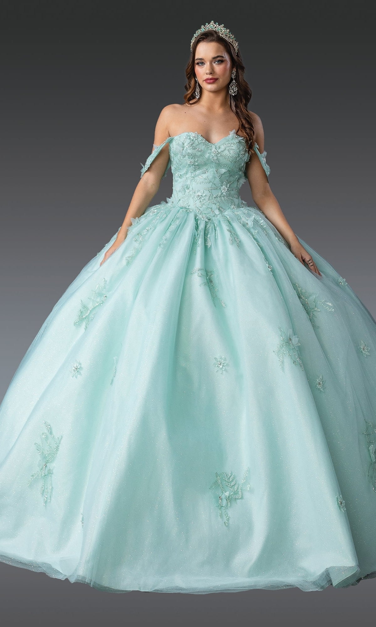 A woman wears the Quinceanera Dress 1922 By Dancing Queen, featuring floral embellishments, a tiara, corset bodice, and glitter tulle ball gown skirt, posed against a gray background.