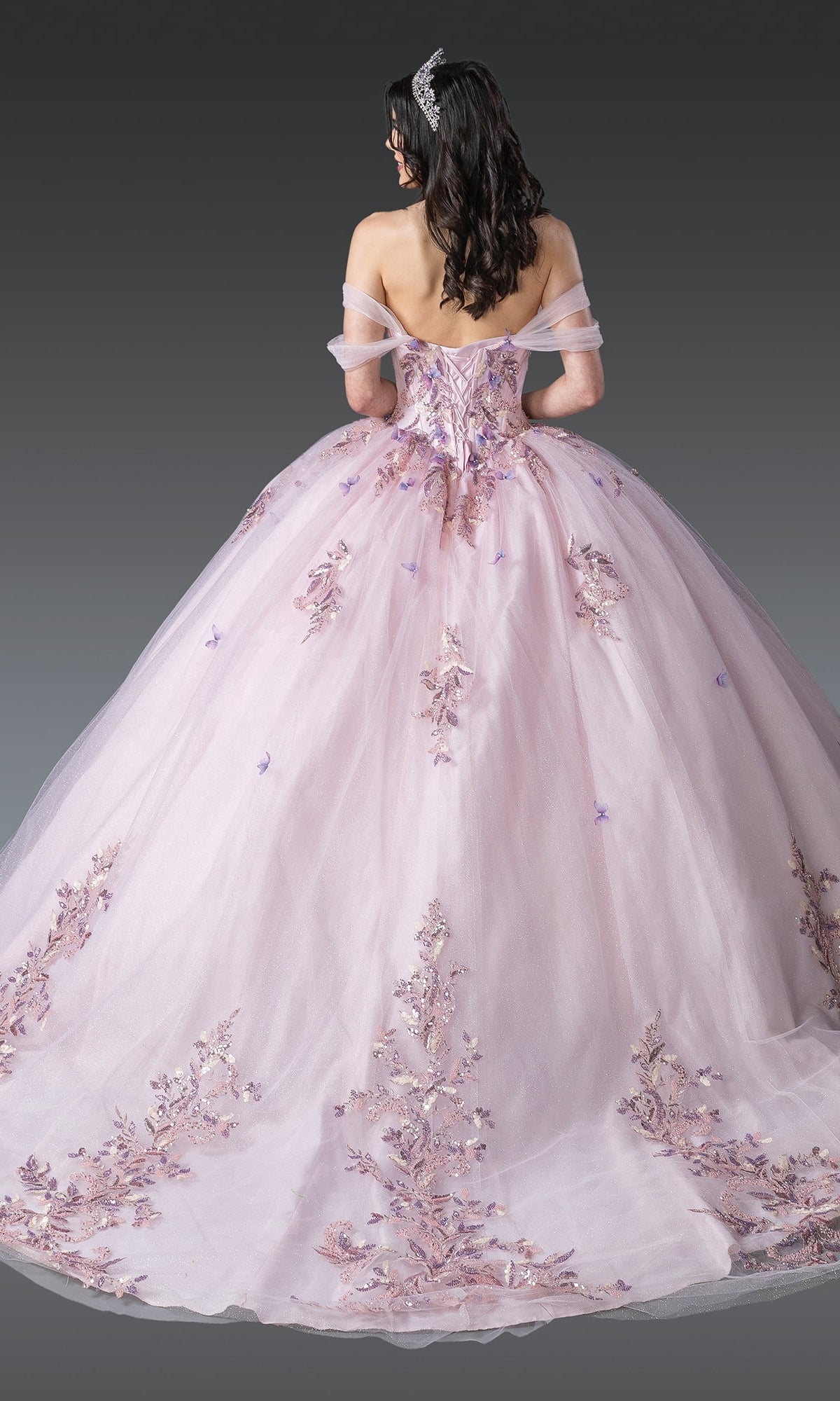 A woman with long dark hair wears the Blush Pink Quinceanera Dress 1924 By Dancing Queen, featuring floral embroidery and a tiara, standing against a plain grey background.
