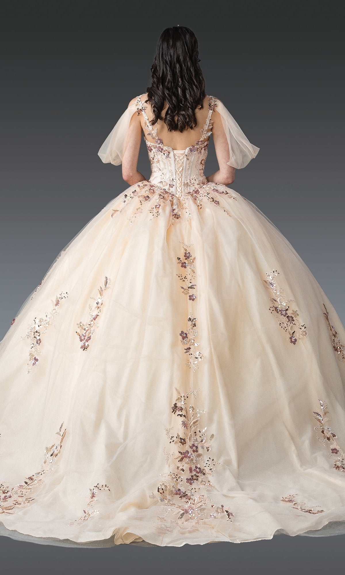 A woman wears the Quinceanera Dress 1926 by Dancing Queen—a cream ball gown featuring sequin lace embroidery, tulle sleeves, and a full skirt—standing with her back to a gray gradient background.