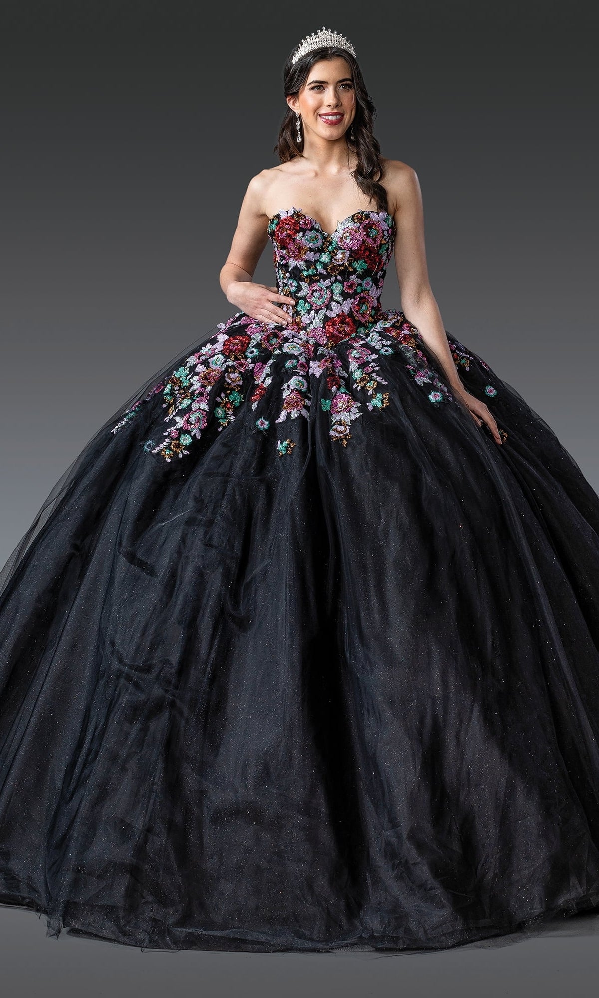 A woman wears the Black Quinceanera Dress 1927 By Dancing Queen, featuring colorful floral embroidery and glitter tulle, standing against a plain gray background with a sparkling tiara completing her look.