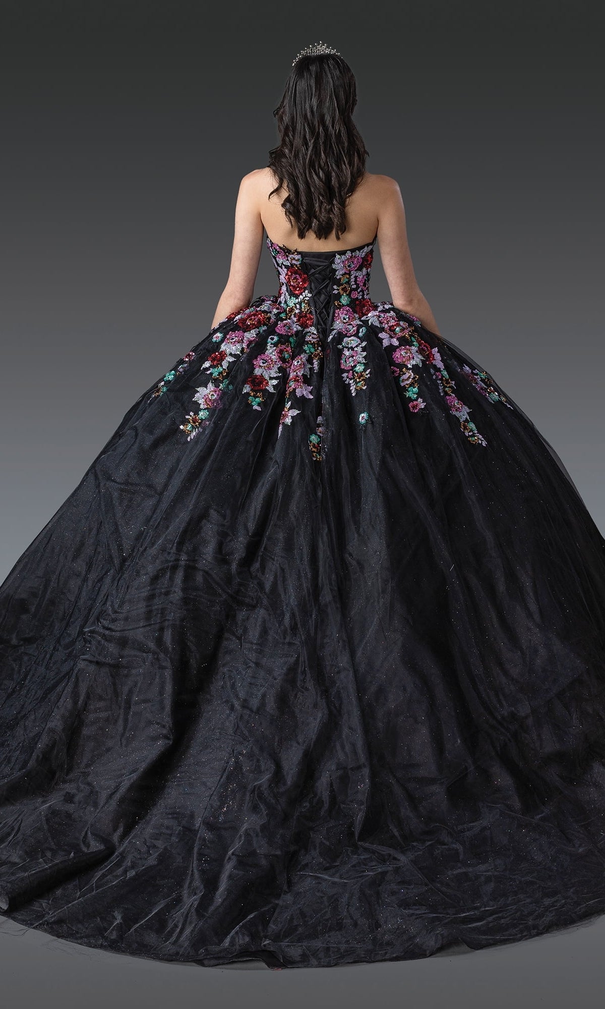 A woman stands facing away, showcasing the full skirt of the Black Quinceanera Dress 1927 By Dancing Queen with colorful floral appliqué and a tiara in her dark hair.