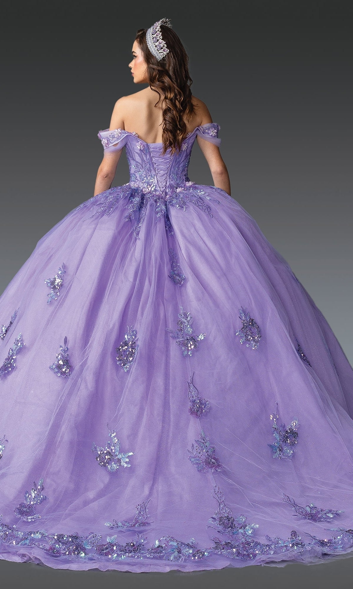 A woman in the Quinceanera Dress 1929 By Dancing Queen, featuring off-the-shoulder lilac design and floral embellishments, wears a tiara and is photographed from the back against a gray background.