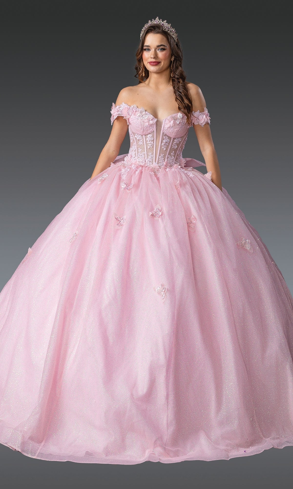 A woman in the Quinceanera Dress 1933 by Dancing Queen, a light pink off-the-shoulder gown with 3D floral appliqués and a tiara, stands against a gray background.