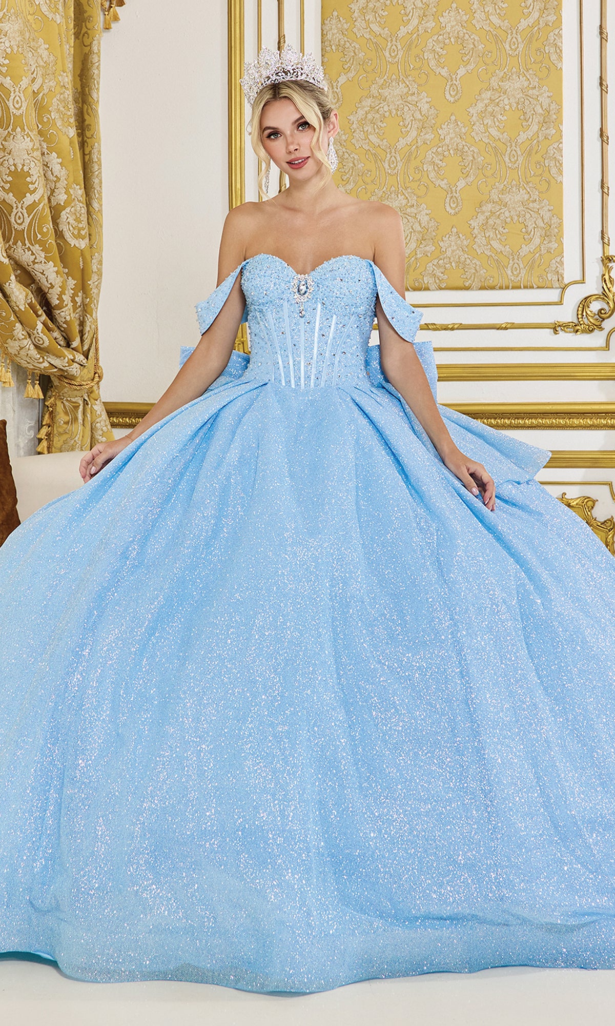 A woman poses in an ornate gold and white room wearing the Quinceanera Dress 1937 By Dancing Queen, a sparkling light blue glitter ball gown with off-the-shoulder straps and a tiara.