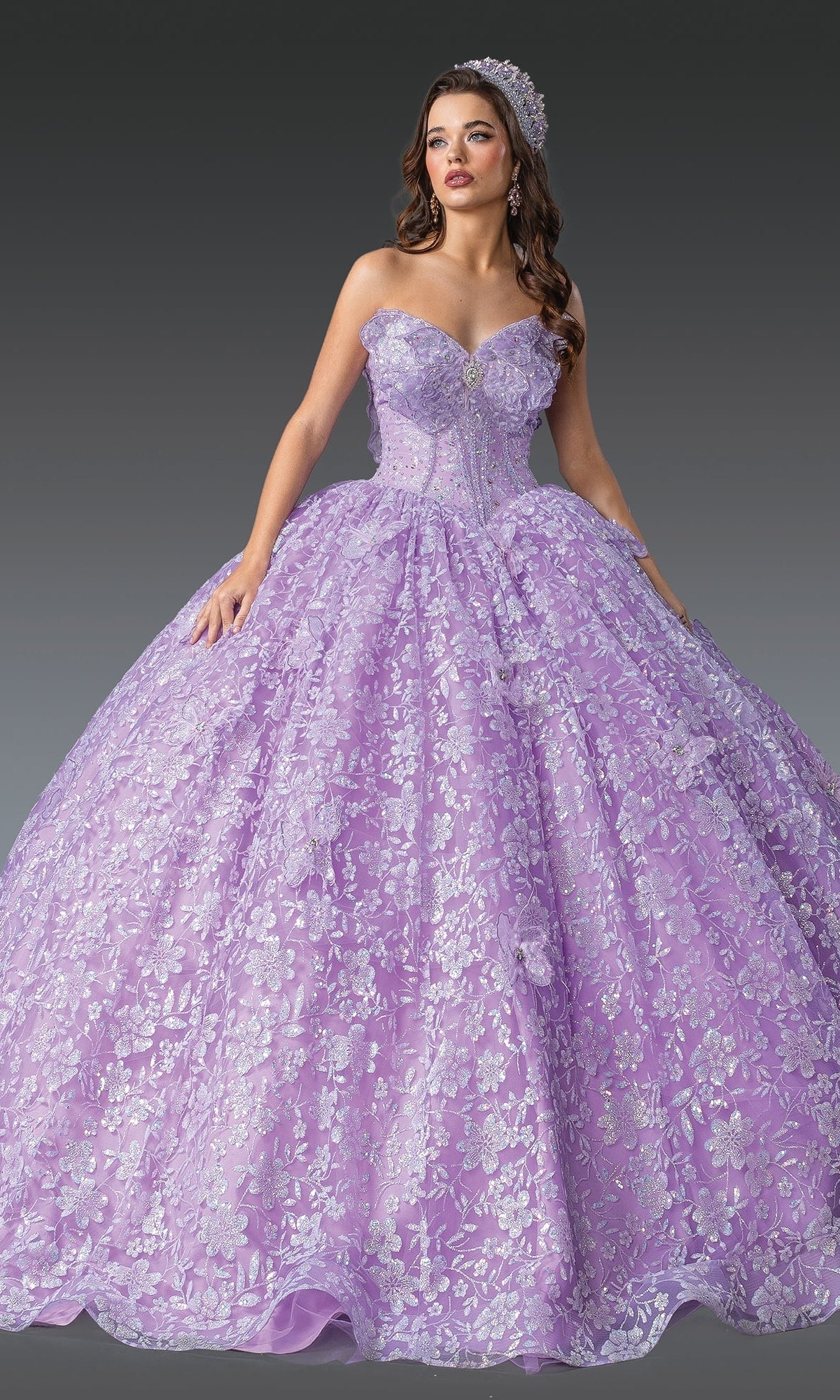 A woman in the Quinceanera Dress 1940 By Dancing Queen—strapless, lilac purple with silver floral details and 3D flowers—wears a jeweled tiara while posing against a gray background.