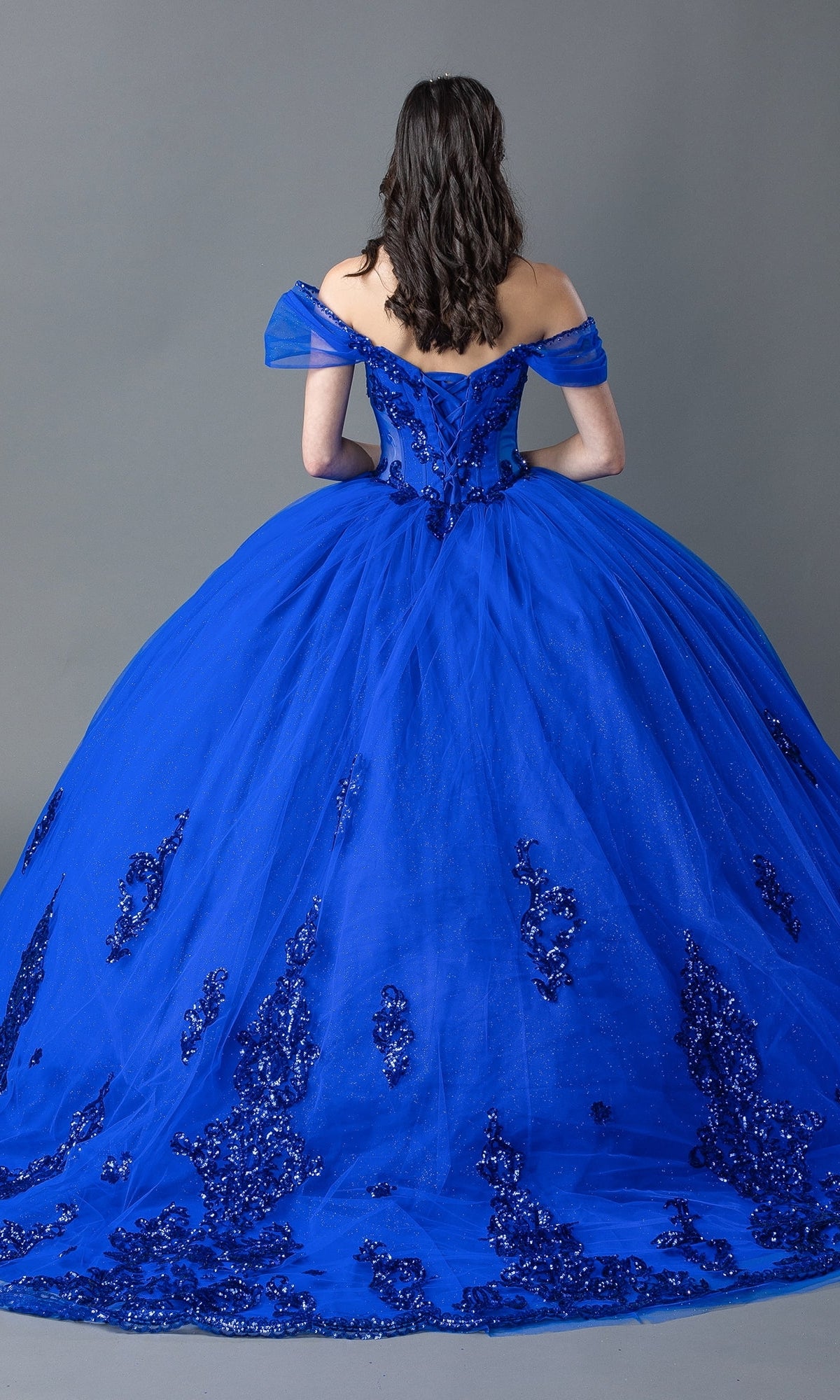 A woman faces away wearing the Quinceanera Dress 1941 By Dancing Queen, an off-the-shoulder royal blue ball gown with a corset bodice, sequin details, and a full tulle skirt.