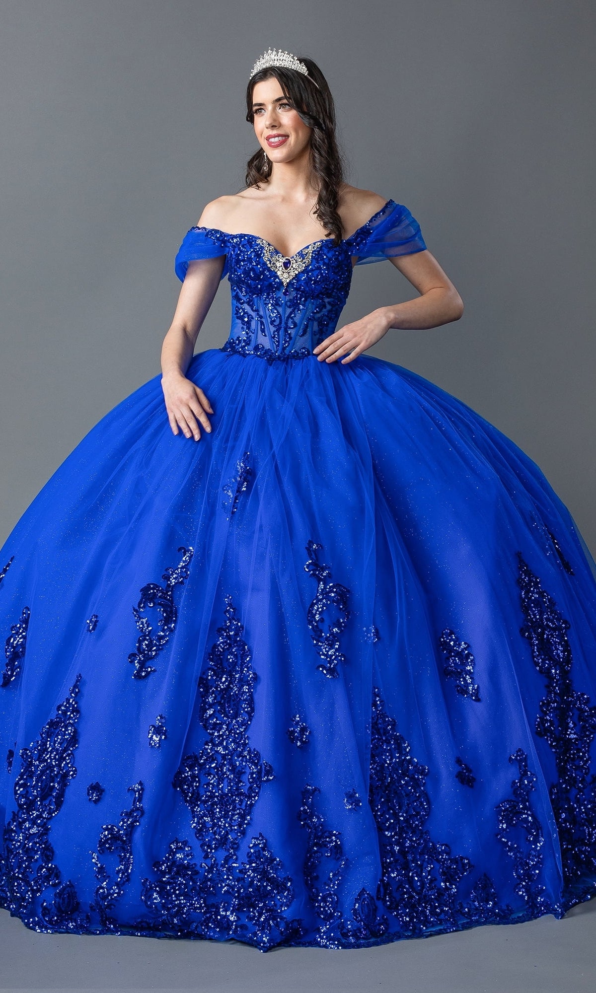 A woman wears the Quinceanera Dress 1941 By Dancing Queen, an off-the-shoulder royal blue ball gown with beaded embroidery and a jeweled tiara, standing against a plain gray background.