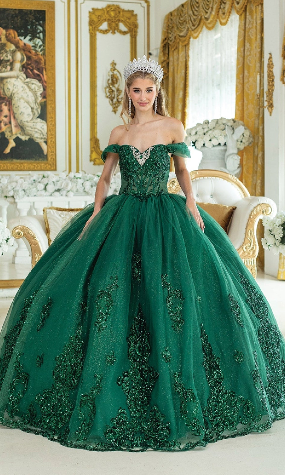 A woman in the Quinceanera Dress 1941 by Dancing Queen—a green, off-the-shoulder ball gown with jeweled beaded embroidery—stands in an ornate room with gold accents and a white sofa.