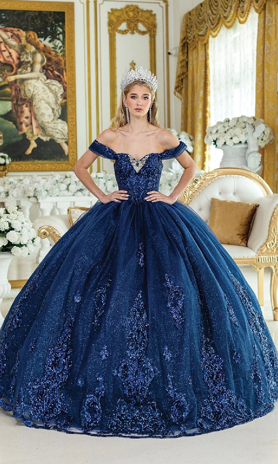 A woman stands in an ornate room with gold decor and white flowers, wearing the Quinceanera Dress 1941 By Dancing Queen—a sparkling blue ball gown featuring a corset bodice and beaded embroidery.