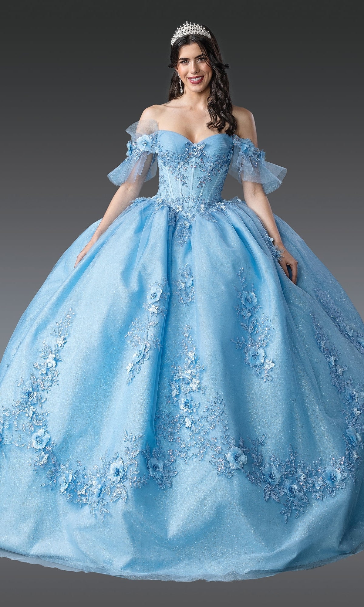 A woman wears the Quinceanera Dress 1944 by Dancing Queen, a blue off-the-shoulder ball gown with floral embroidery and corset bodice, paired with a tiara, standing against a plain gray background.