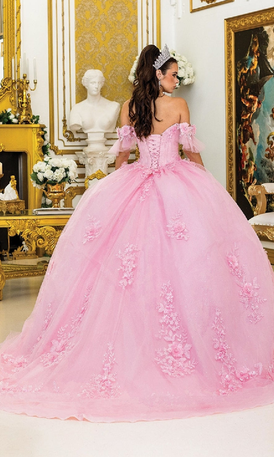 A woman wearing the Quinceanera Dress 1944 By Dancing Queen, featuring pink fabric and floral details, stands in an ornate room with a tiara, facing away from the camera.