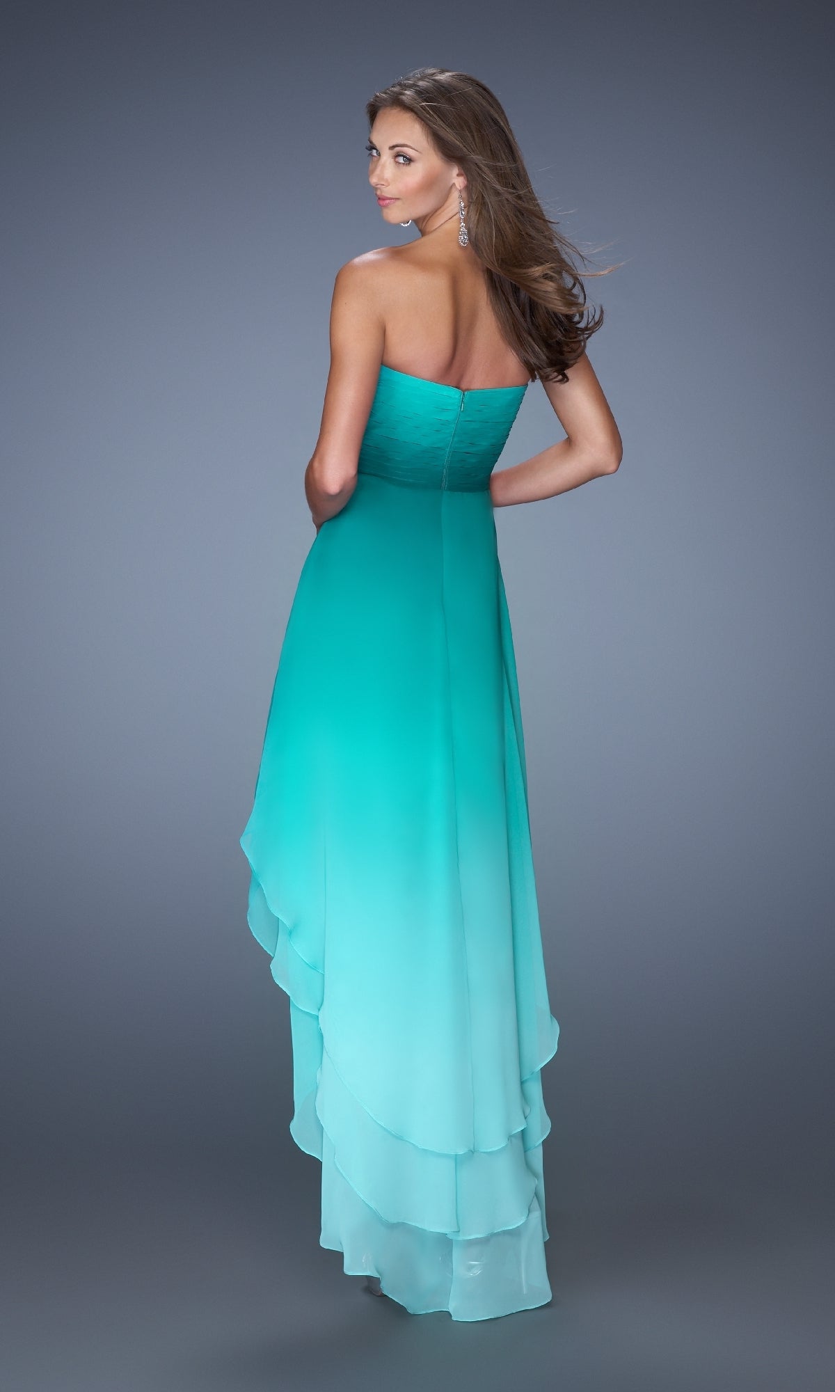 A woman stands with her back to the camera, showcasing the High-Low Prom Dress: La Femme 19467—a strapless gown with layered turquoise ombre hues—against a plain gray background.