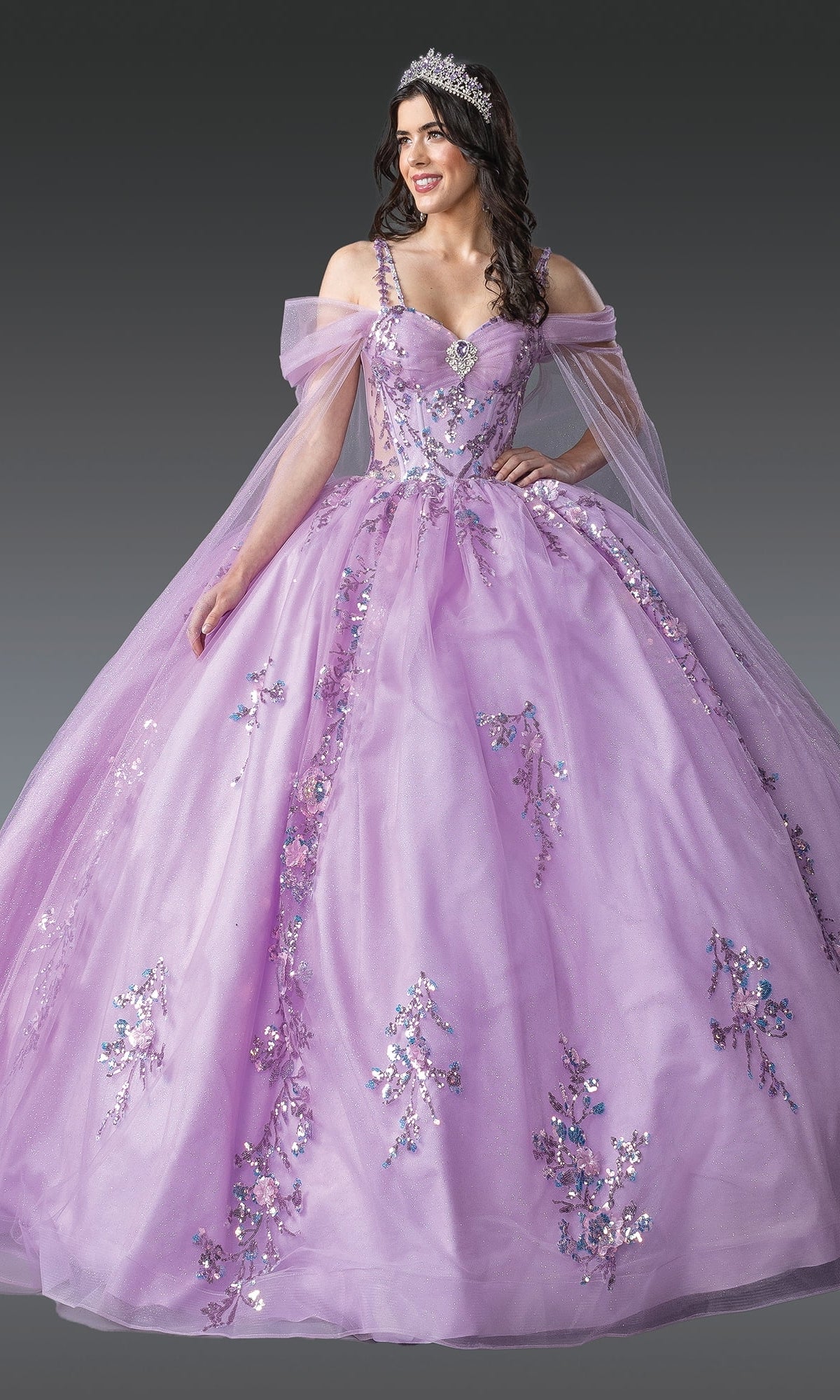 A woman wears the Quinceanera Dress 1947 by Dancing Queen, featuring lavender lace embroidery and floral details, paired with a tiara, standing against a gray background.