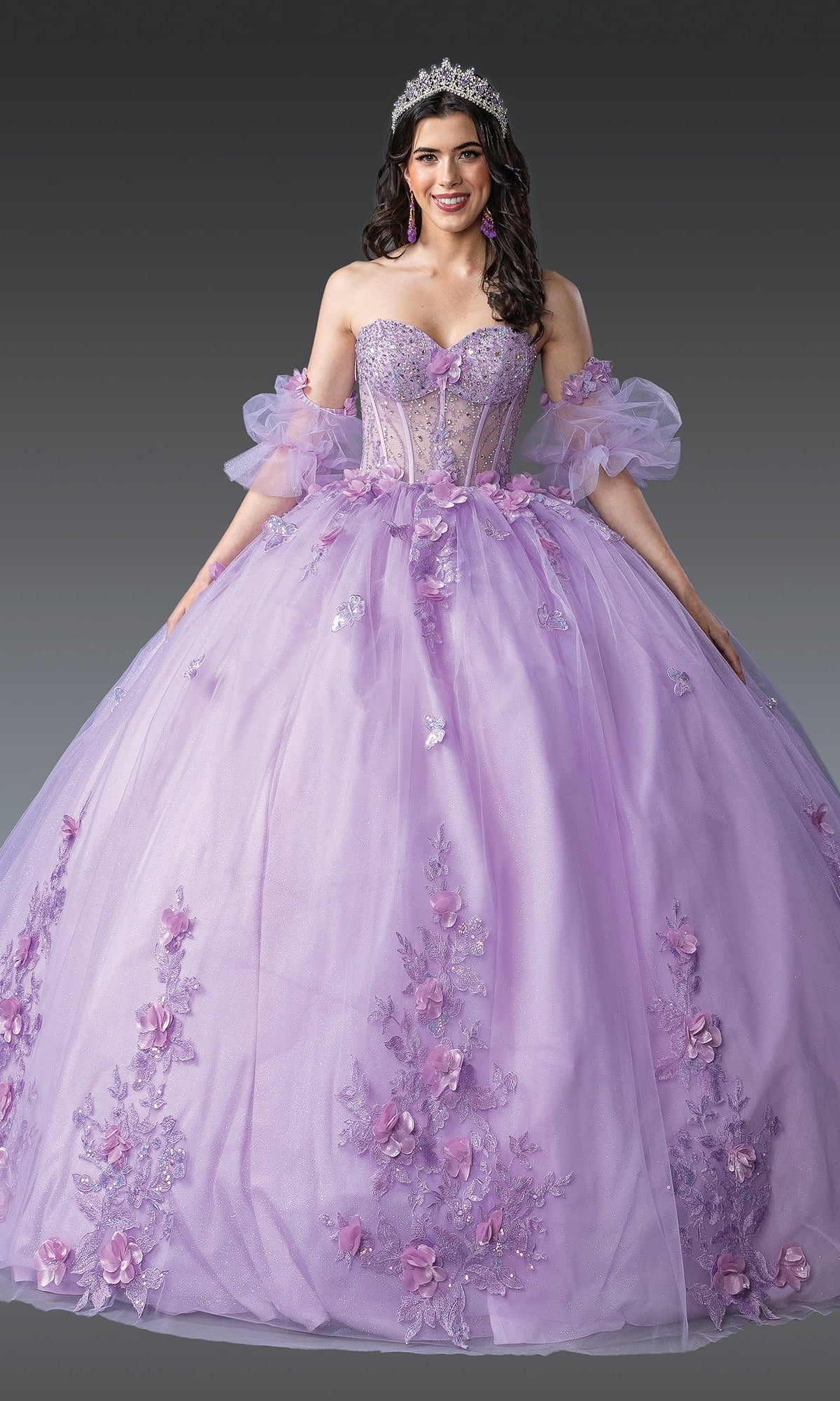 A woman wears the Quinceanera Dress 1949 By Dancing Queen—lilac glitter tulle with floral appliqués and puffed sleeves—plus a tiara and earrings, standing against a gray background.