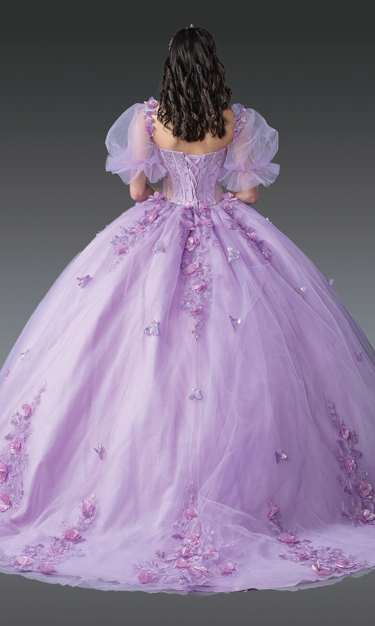 A woman with dark wavy hair faces away, wearing the Quinceanera Dress 1949 By Dancing Queen—a floor-length lavender glitter tulle ball gown with floral appliqués and tulle puff sleeves.