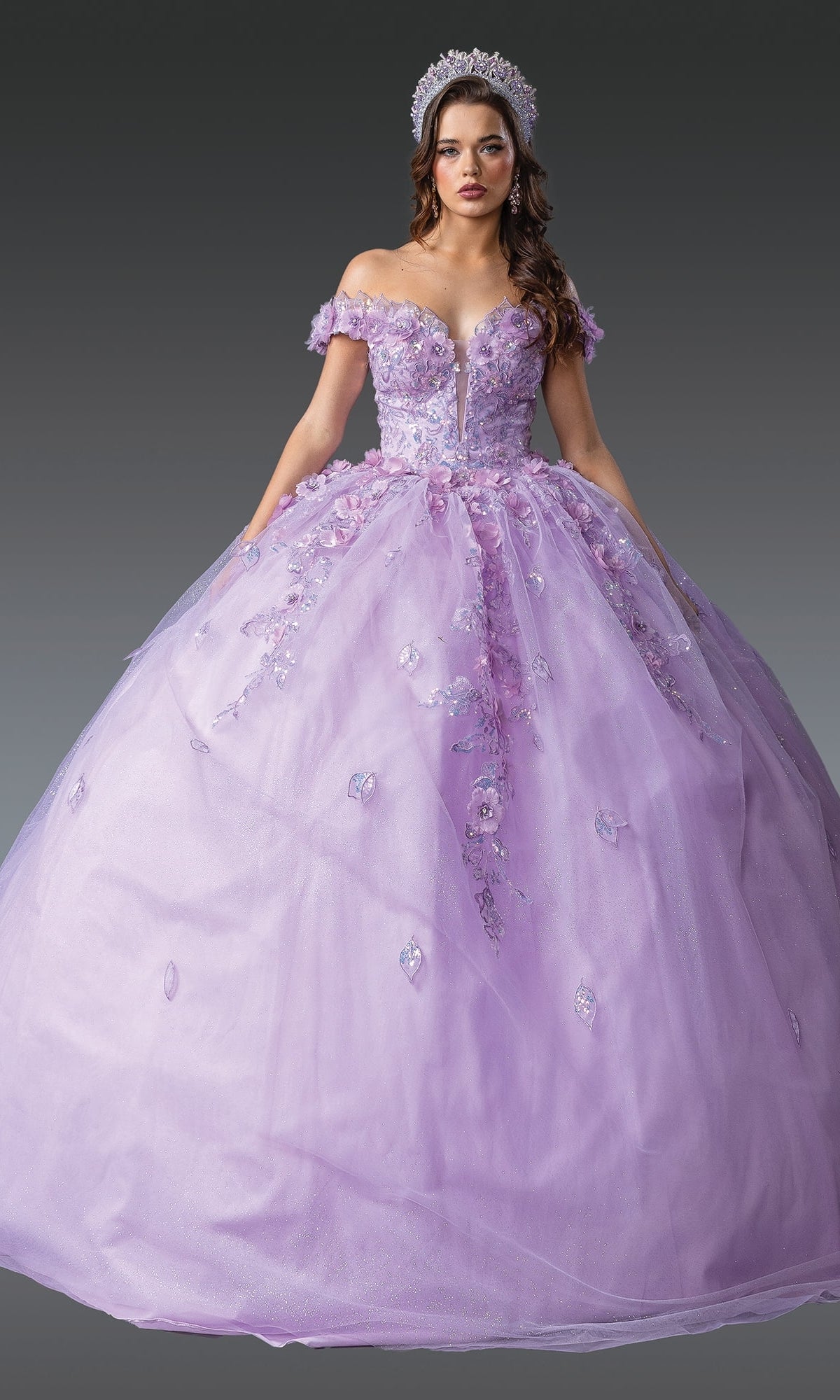 A young woman wears the Quinceanera Dress 1951 By Dancing Queen, featuring an off-the-shoulder lilac design with floral appliqué and a ball gown skirt, paired with a jeweled tiara, set against a plain gray background.