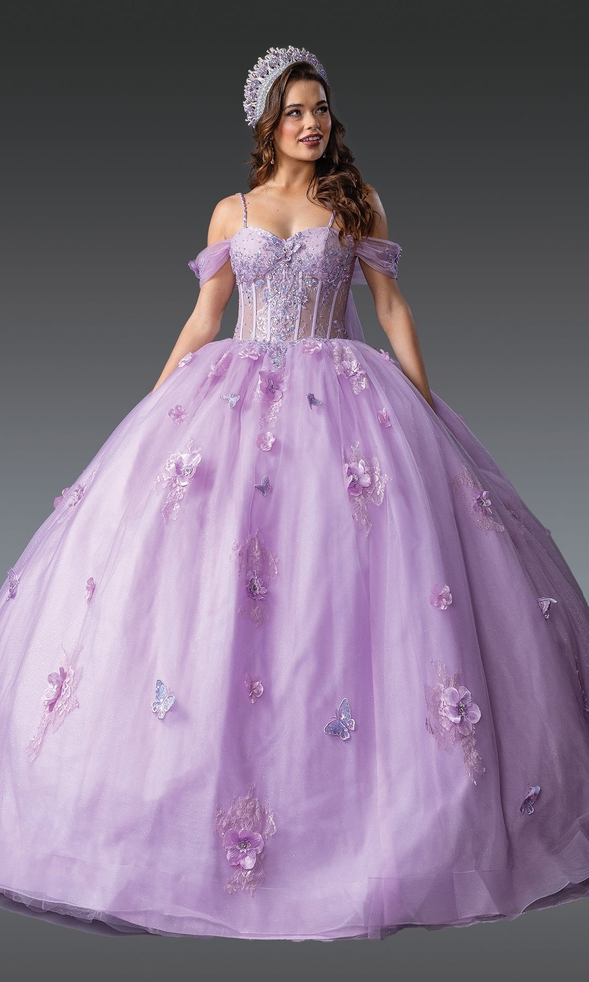 A woman in Quinceanera Dress 1954 By Dancing Queen—with a lavender lace corset bodice, floral and butterfly accents, and matching headpiece—stands before a gray background.