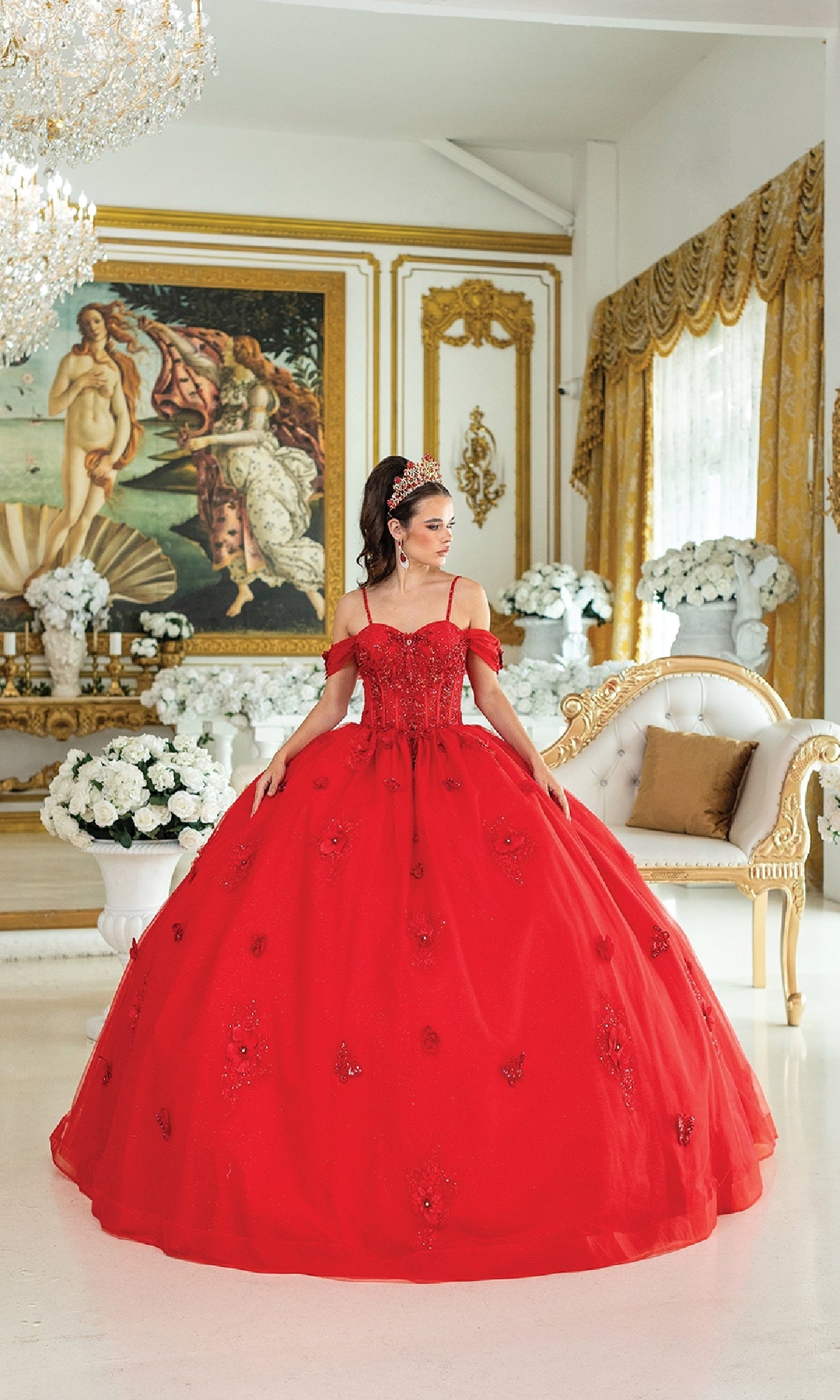 A woman wearing the Quinceanera Dress 1954 By Dancing Queen, a glitter tulle ball gown with a lace corset bodice, stands in an ornate room decorated with gold accents, white flowers, and a classical painting.