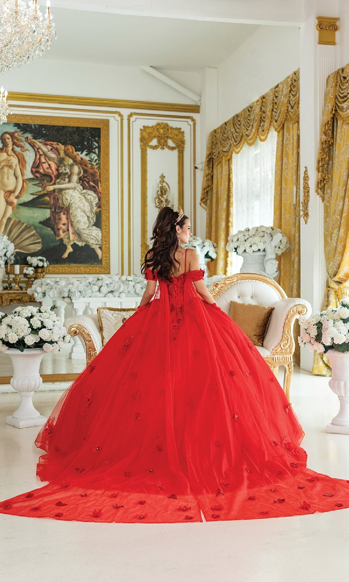 Wearing the Quinceanera Dress 1954 By Dancing Queen, a woman sits on a cream sofa in an ornate gold room adorned with white flowers and classical art.