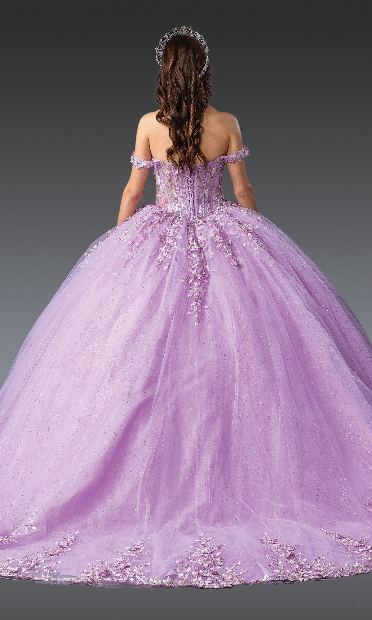 A woman with long brown hair wears the Quinceanera Dress 1955 By Dancing Queen, an off-the-shoulder lavender ball gown with floral lace embroidery and a matching headpiece, seen from behind against a gray background.