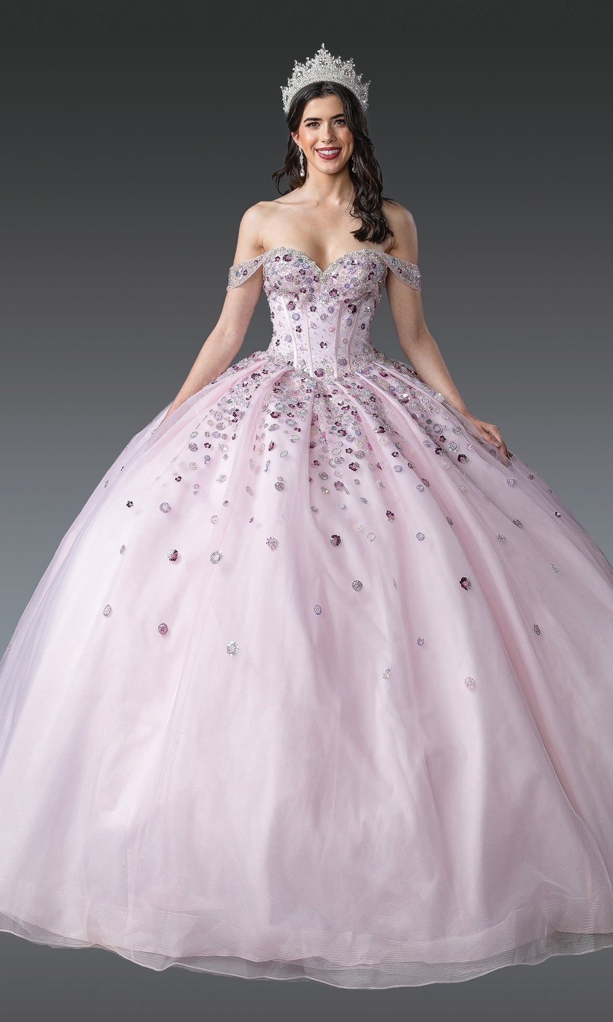 A woman stands against a plain gray background wearing the Quinceanera Dress 1959 By Dancing Queen—an off-shoulder blush pink gown with jeweled detailing, full ball skirt, corset bodice, and a silver crown.