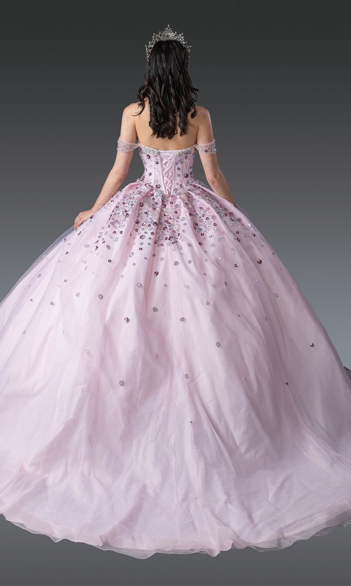 Woman in the Quinceanera Dress 1959 By Dancing Queen, a blush pink ball gown with off-the-shoulder sleeves, jeweled accents, and corset bodice, wearing a tiara, seen from the back against a gradient background.