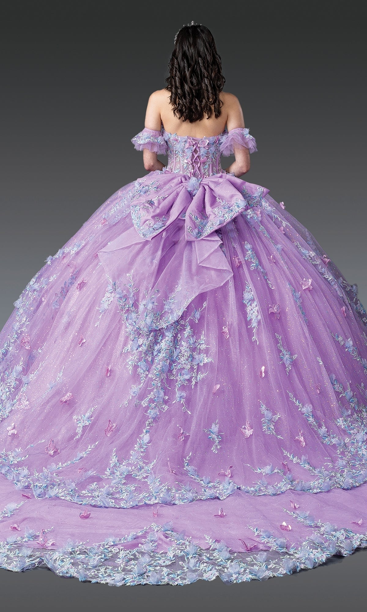 A person wears the Quinceanera Dress 1961 By Dancing Queen, featuring lilac purple fabric, a corset bodice, floral embellishments, a large bow, and a flowing decorated train, standing against a plain dark background.