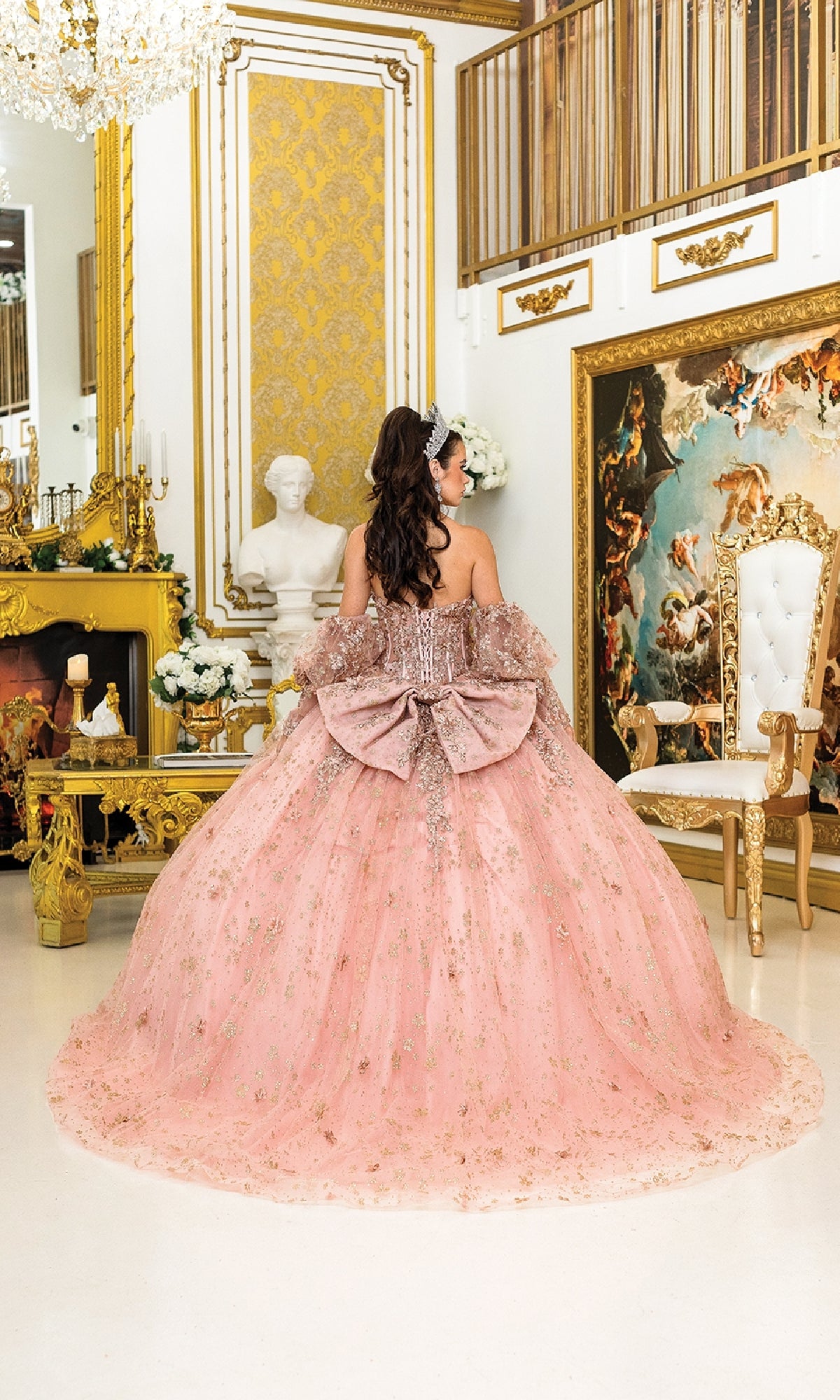 A young woman in the Quinceanera Dress 1966 by Dancing Queen, featuring glitter details and a large bow, stands in an ornate room with gold accents, her back turned to the camera.