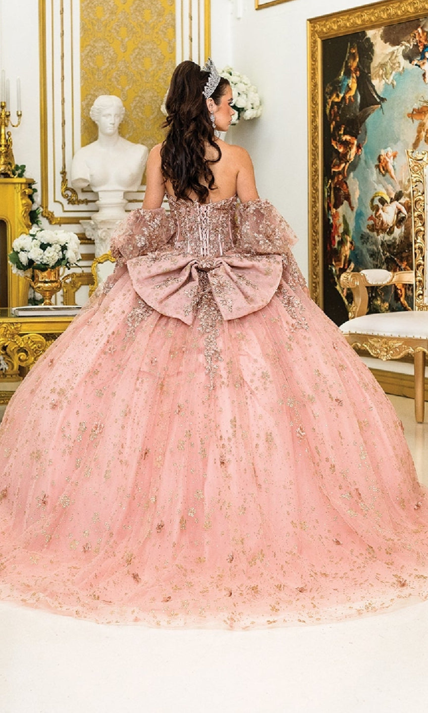 A young woman in the Quinceanera Dress 1966 by Dancing Queen, featuring a large bow, stands in a lavish room adorned with gold accents, flowers, a statue, and a grand painting.