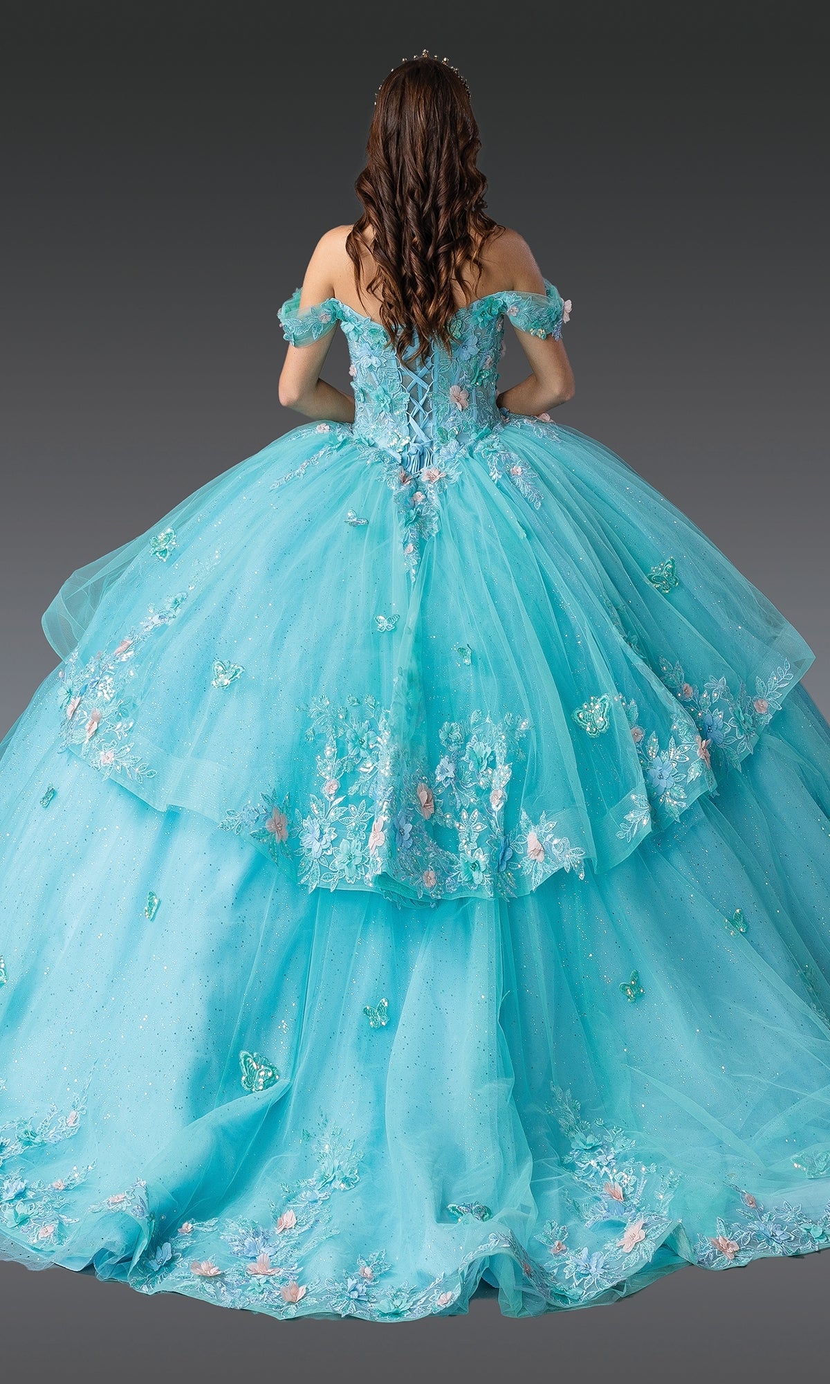 A person with long, wavy hair stands facing away, wearing the voluminous Quinceanera Dress 1974 By Dancing Queen in blue with floral embroidery and butterfly decorations, set against a gray background.