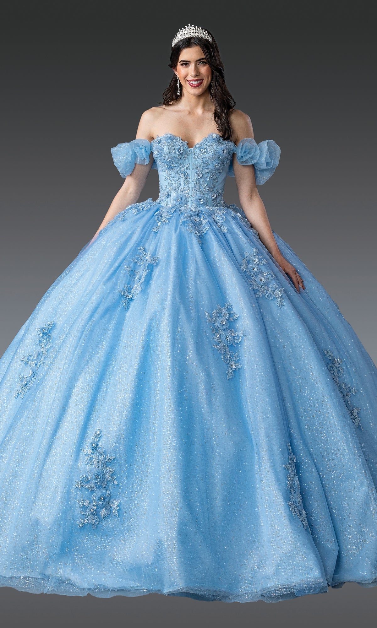 A woman in the Quinceanera Dress 1976 By Dancing Queen, featuring a blue ball gown skirt, corset bodice, and floral embellishments, poses with a tiara against a plain gray background.