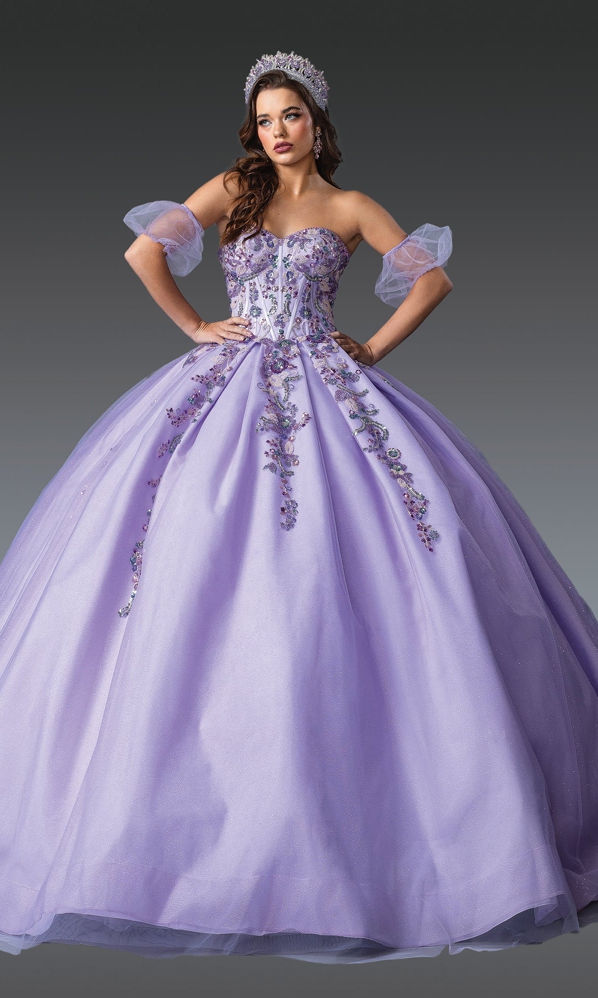 Woman wearing the Lilac Quinceanera Dress 1977 by Dancing Queen, featuring purple glitter tulle, floral and beaded embroidery, sheer puff sleeves, and a jeweled tiara—perfect for a modern quinceanera look against a gray background.