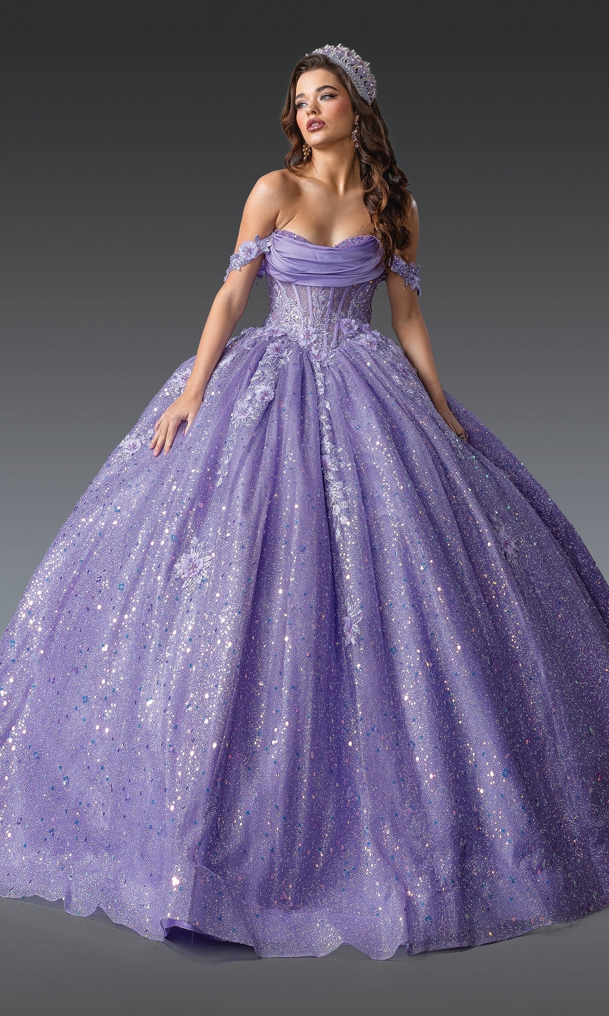A woman stands against a gray background wearing the Quinceanera Dress 1984 by Dancing Queen, an off-the-shoulder sparkling lavender ball gown paired with a matching tiara.
