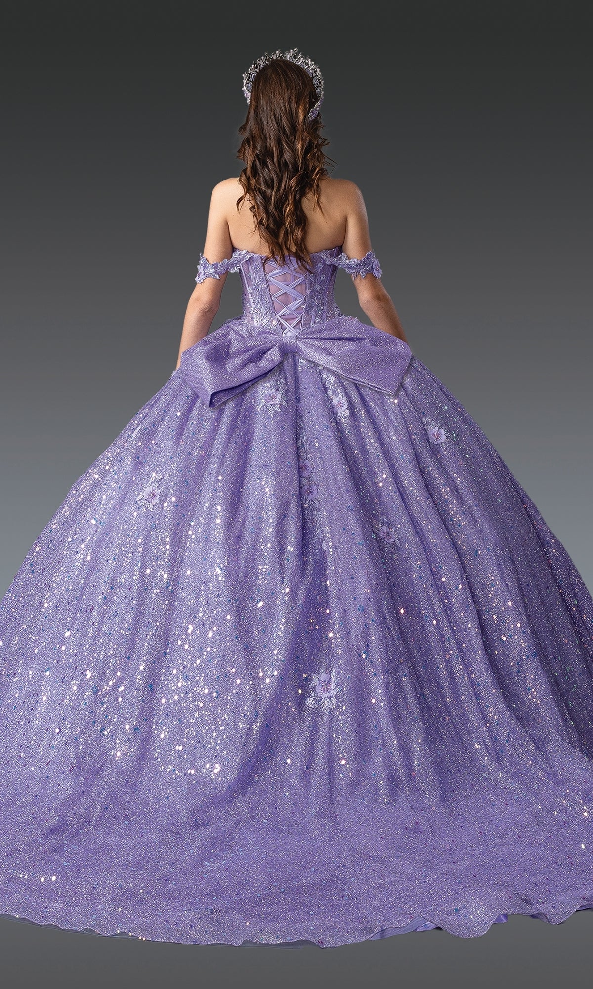A woman with long brown hair, facing away from the camera against a gray background, wears Quinceanera Dress 1984 by Dancing Queen—a sparkly lavender gown with an off-the-shoulder neckline and a large back bow.