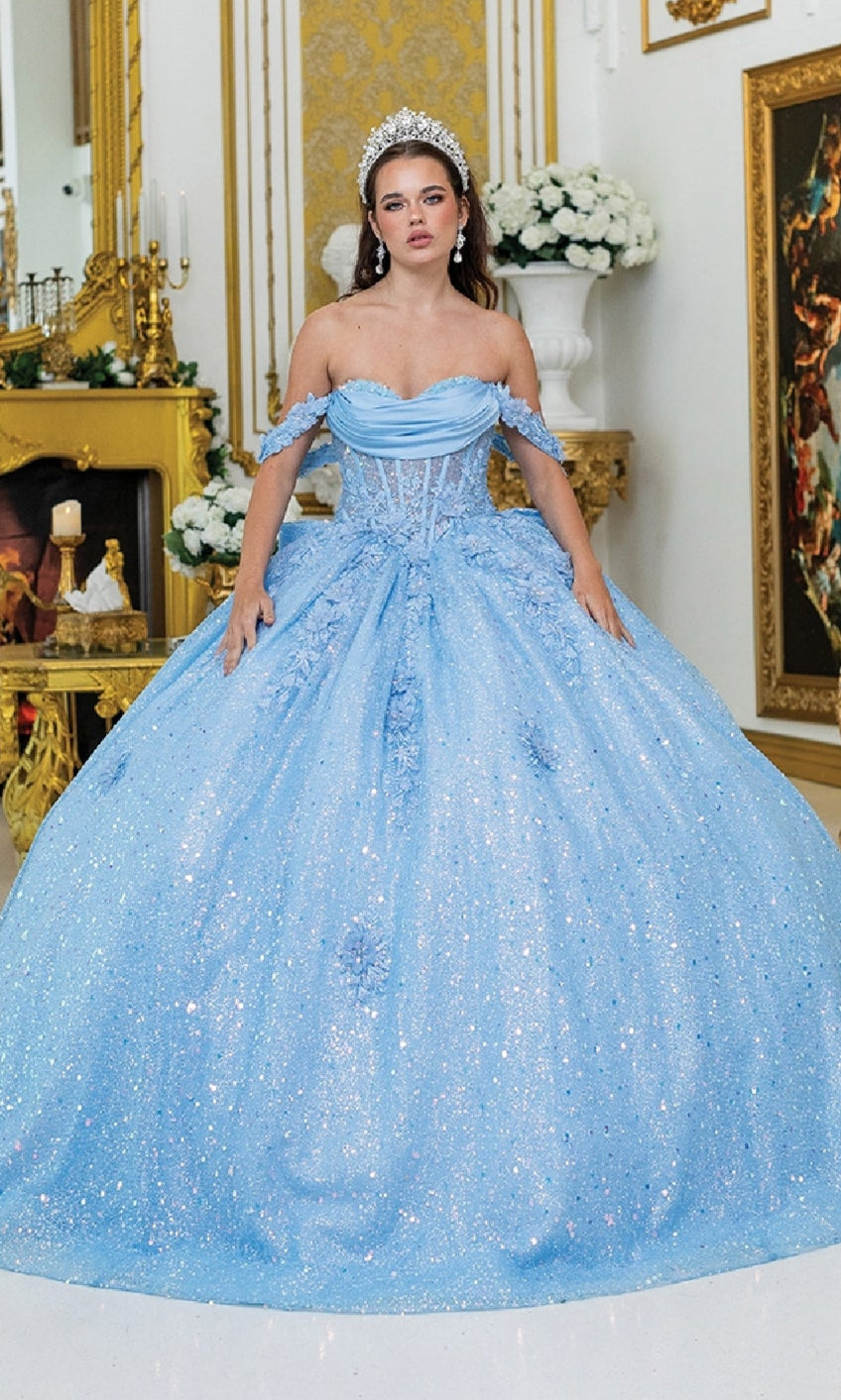 A woman wears the Quinceanera Dress 1984 by Dancing Queen, a sparkling light blue off-the-shoulder ball gown, with a tiara, standing indoors in an ornate room adorned with gold accents and white flowers.