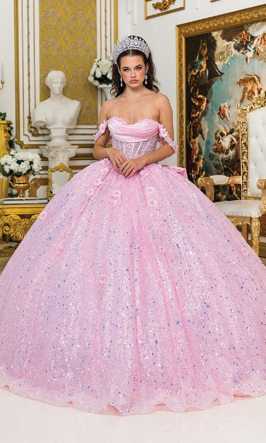 A woman in the Quinceanera Dress 1984 By Dancing Queen, featuring an off-the-shoulder pink sparkling ball gown, stands in an ornate room with gold accents, a bust statue, and classical artwork in the background.