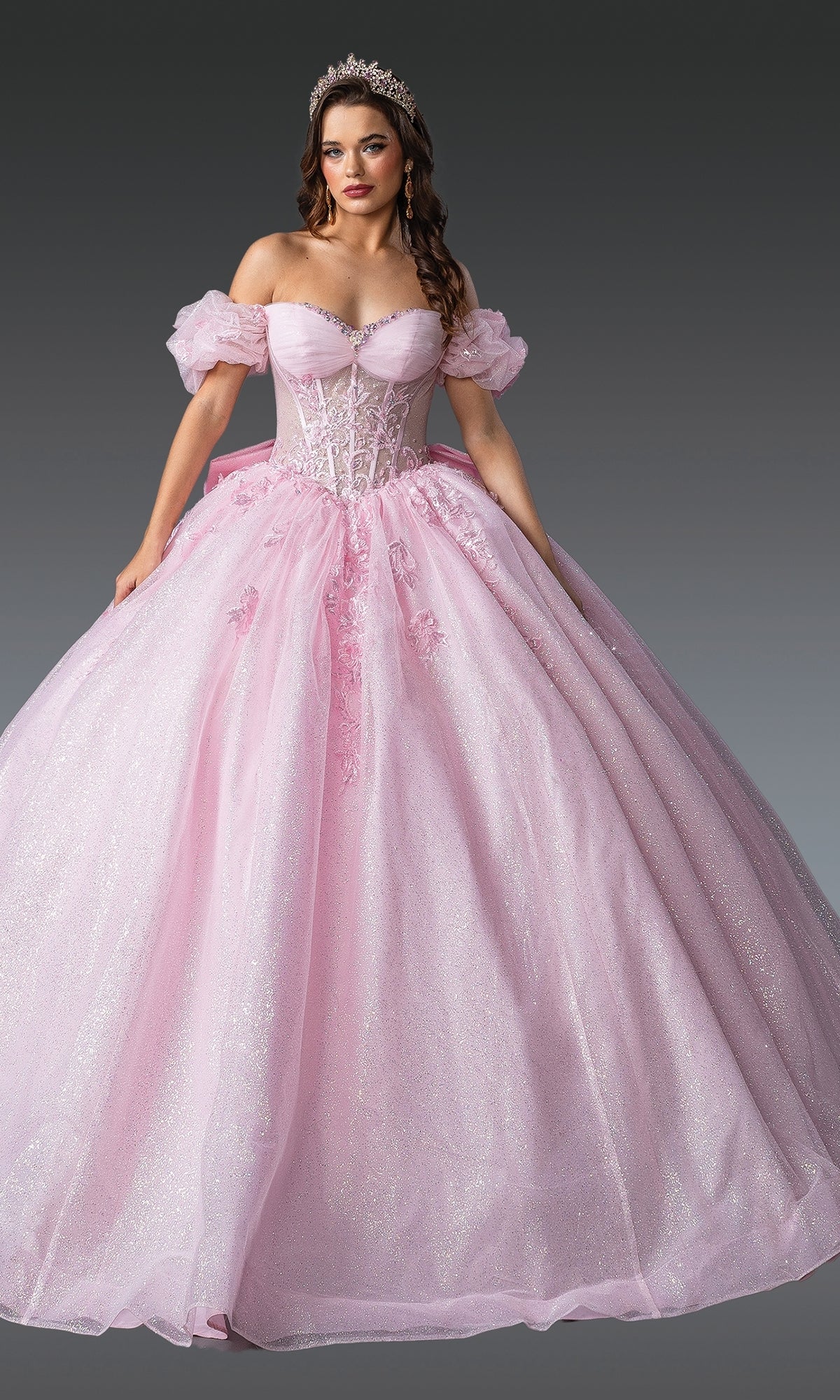 A woman in the Quinceanera Dress 1985 By Dancing Queen, featuring a sparkling pink ball gown with a lace embroidered corset bodice and off-the-shoulder sleeves, poses with a tiara against a neutral gray background.