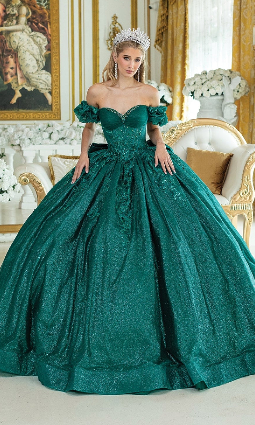 A woman wearing the Quinceanera Dress 1985 By Dancing Queen, a sparkling emerald green ball gown with a corset bodice and tiara, stands in an ornate room adorned with gold decor, white flowers, and elegant furniture.