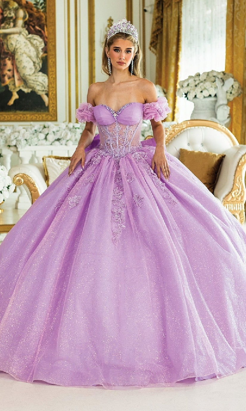 A woman in the Quinceanera Dress 1985 By Dancing Queen, a sparkling lavender ball gown with a corset bodice and tiara, stands in an ornate room adorned with gold accents and white floral arrangements.