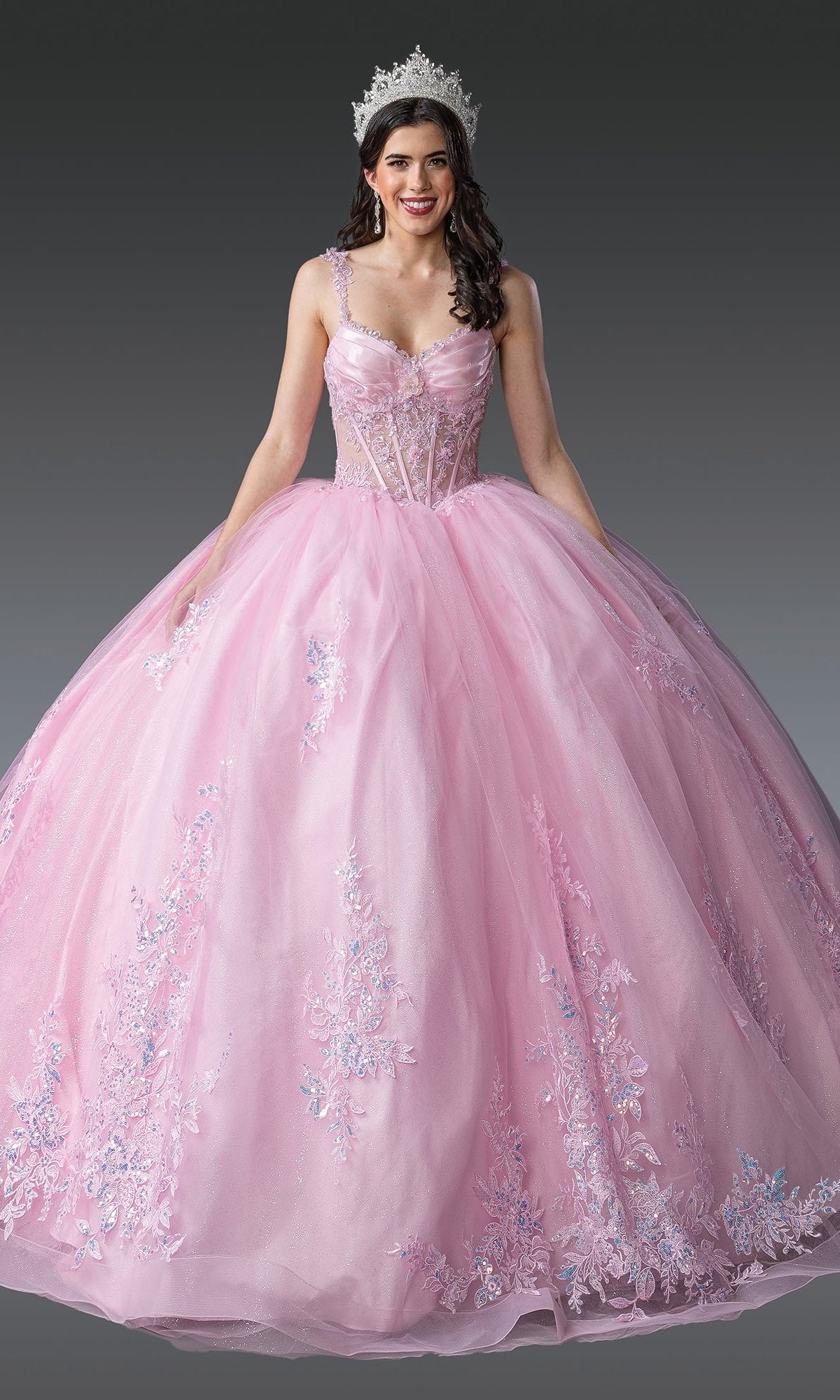 Woman wearing the Quinceanera Dress 1989 by Dancing Queen, a pink ball gown with floral and beaded embroidery and a tiara, standing against a plain gray background.