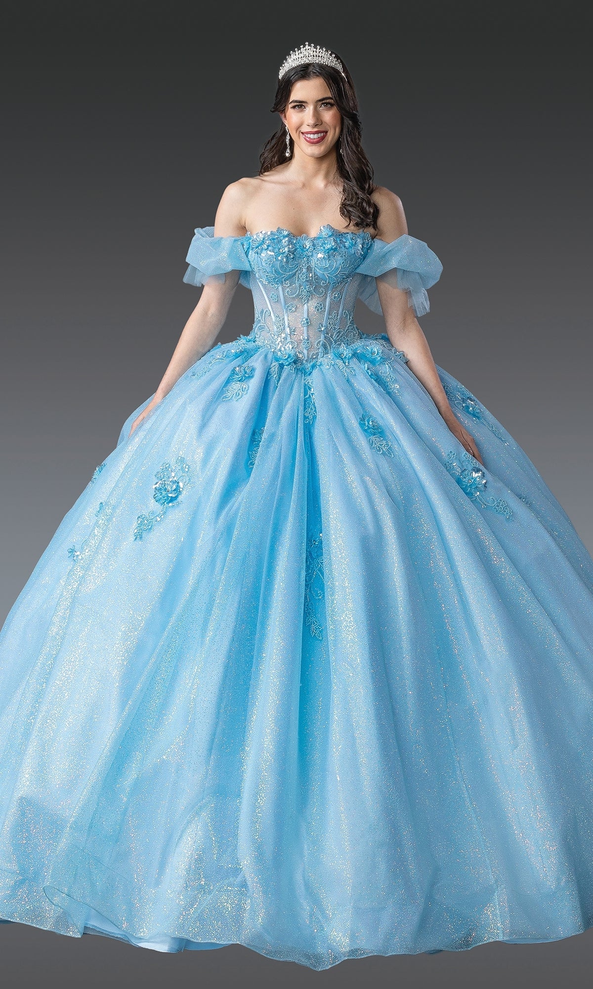 A woman wears the Quinceanera Dress 1990 By Dancing Queen—a sparkling light blue off-the-shoulder ball gown with a corset bodice and tiara—standing against a plain gray background.