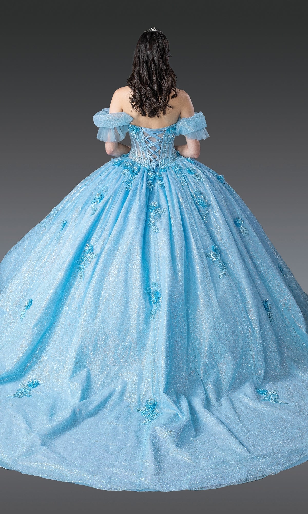 A woman faces away, revealing the corset laced-back and full ruffled tulle skirt of the Dancing Queen Quinceanera Dress 1990—an off-the-shoulder light blue ball gown adorned with floral details.
