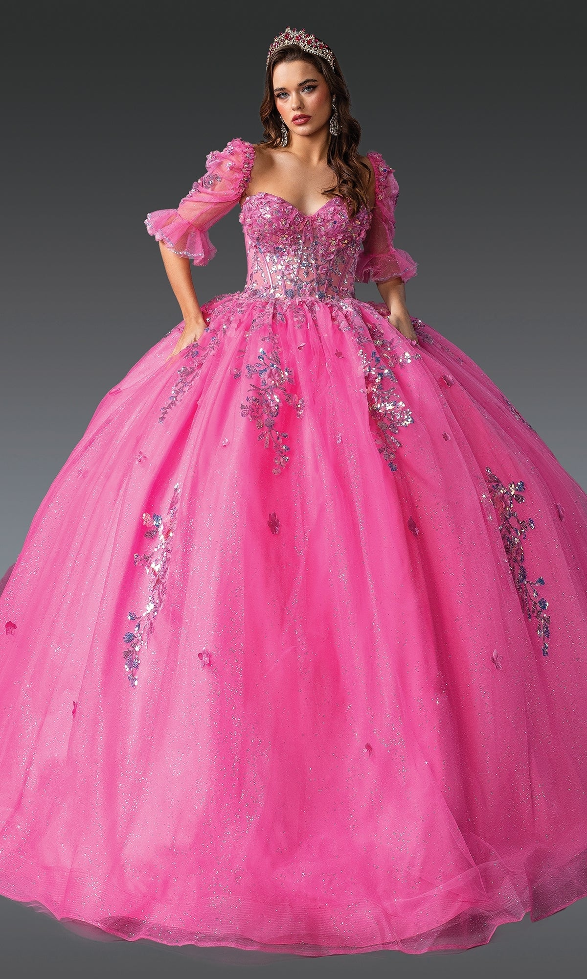 Woman wearing the Pink Quinceanera Dress 1995 By Dancing Queen—bright pink tulle ball gown with sequin floral embroidery and puffed sleeves—stands against a plain gray background, topped with a tiara.