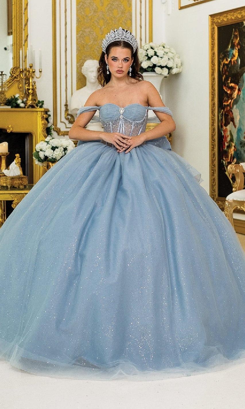 A woman wearing the Quinceanera Dress 1996 By Dancing Queen, a sparkling light blue gown with glitter tulle and a tiara, stands indoors amid ornate gold décor and white flowers.